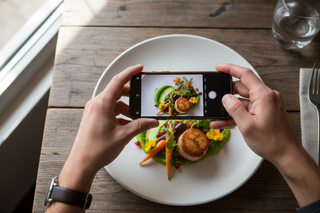 Two hands holding a phone, over a plate of food. The angle shows a birds eye view looking through the phone and the plate beneath it. o