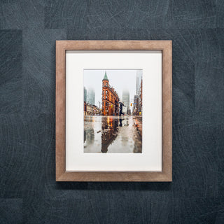 Framed photograph of a cityscape with a couple walking on a rainy day, displayed on a textured grey wall in a bronze frame